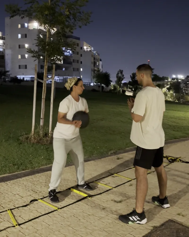 המאמן מלווה מתאמנת מקבוצת האמהות בתרגיל כדור כוח על סולם זריזות בפארק